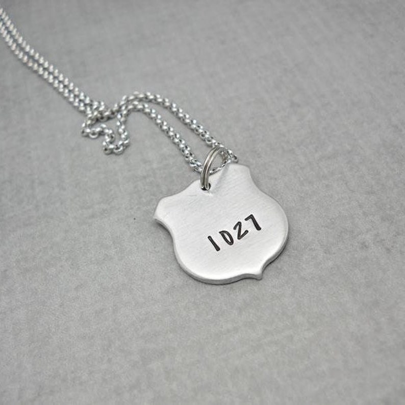 May include: A silver chain necklace with a silver shield pendant. The shield has the number "1027" engraved on it.
