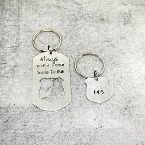 May include: Two silver keychains with a police badge design. One keychain has the text "Always come home safe to me" engraved on it. The other keychain has the number "143" engraved on it.