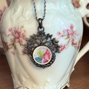 May include: A silver-toned necklace featuring a circular pendant with a floral ceramic inlay. The pendant is framed by a metal tree-of-life design. The necklace is draped over a cream-colored porcelain creamer with pink floral patterns.