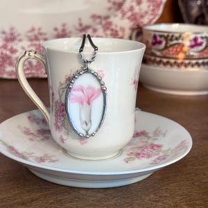 May include: An oval pendant featuring a pink flower illustration on a white background, encased in a silver-toned metal frame with a beaded edge, suspended from a thin black cord, resting against a cream-colored teacup with pink floral patterns.