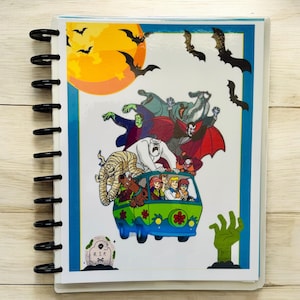 May include: A spiral-bound notebook featuring a colorful illustration of a cartoon scene with a green van, characters, and bats against a yellow moon. The notebook has a blue border and a wooden background.