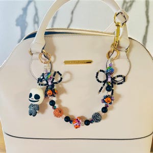 Puede incluir: Un bolso crema con detalles dorados y un charm decorativo de cuentas. El charm presenta una figura de Jack Skellington, lazos negros y cuentas coloridas con acentos de calabaza y flores. El bolso es de la marca Steve Madden.
