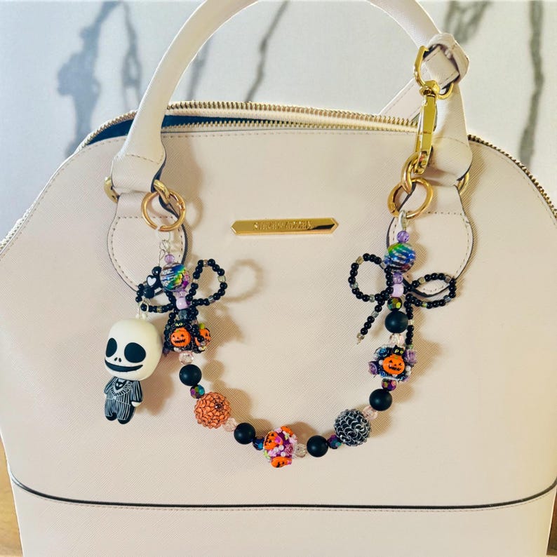 Puede incluir: Un bolso crema con detalles dorados y un charm decorativo de cuentas. El charm presenta una figura de Jack Skellington, lazos negros y cuentas coloridas con acentos de calabaza y flores. El bolso es de la marca Steve Madden.