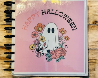 May include: A planner with a pink cover featuring a white ghost illustration surrounded by flowers. The words "HAPPY HALLOWEEN" are arched above the ghost. The planner has black binding rings.