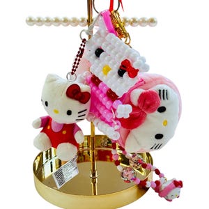 May include: A collection of Hello Kitty-themed keychains and accessories. Includes a plush Hello Kitty, a beaded keychain, and a pink heart-shaped item. The items are displayed on a gold-colored stand with a pearl-like accent.
