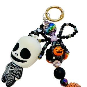 Puede incluir: Un llavero tem&aacute;tico de Halloween con una figura de Jack Skellington y una ara&ntilde;a de cuentas con una calabaza. El llavero incluye un cierre dorado y varias cuentas en colores negro, naranja e iridiscente. La figura de Jack Skellington es blanca con detalles negros.