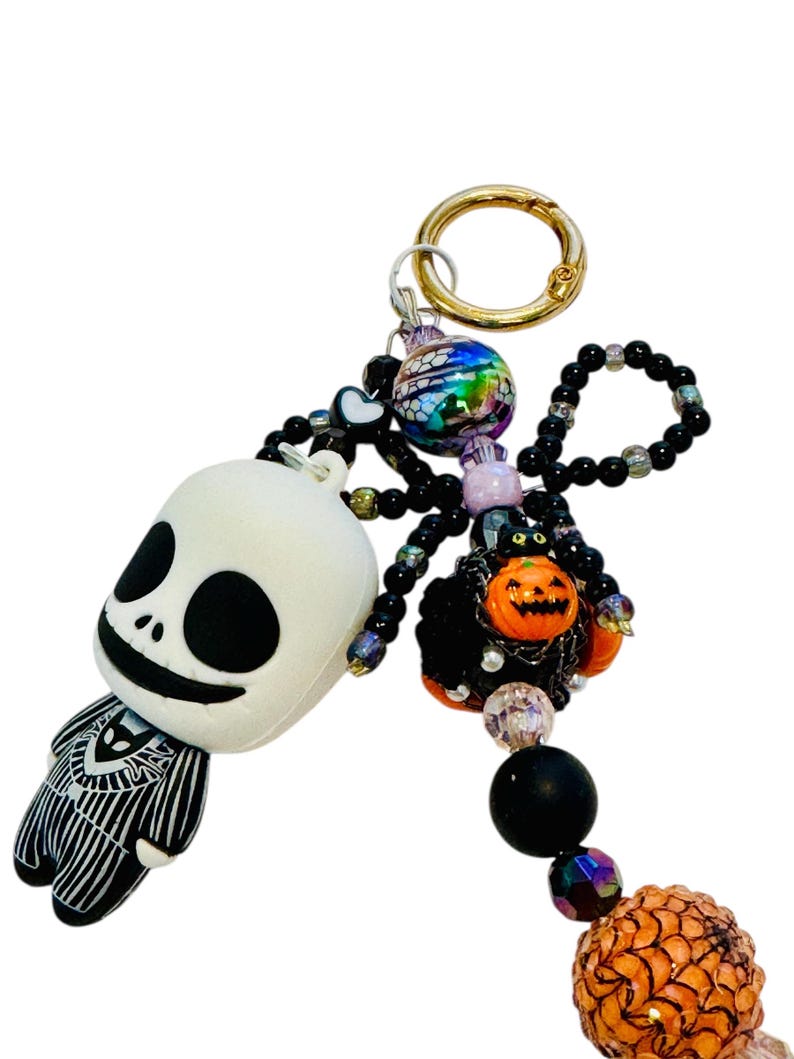 Puede incluir: Un llavero tem&aacute;tico de Halloween con una figura de Jack Skellington y una ara&ntilde;a de cuentas con una calabaza. El llavero incluye un cierre dorado y varias cuentas en colores negro, naranja e iridiscente. La figura de Jack Skellington es blanca con detalles negros.
