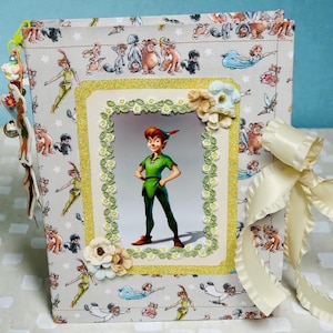 May include: A handmade scrapbook featuring Peter Pan. The cover is decorated with a Peter Pan image in a gold glitter frame, surrounded by floral embellishments. The background fabric has a repeating pattern of Peter Pan characters.