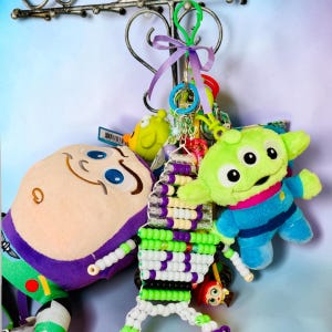 May include: A collection of keychains featuring a plush Buzz Lightyear character, a plush three-eyed green alien, and a beaded figure made of white, purple, and green plastic pony beads, all attached to a green plastic clip with a purple ribbon.