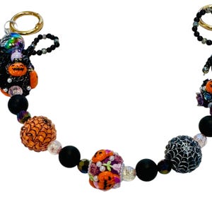 Puede incluir: Un llavero de cuentas con tem&aacute;tica de Halloween con anillos dorados. Las cuentas incluyen cuentas con forma de calabaza naranja, cuentas negras mate y una cuenta con un dise&ntilde;o de telara&ntilde;a. El llavero tambi&eacute;n presenta una figura negra con detalles de calabaza naranja.
