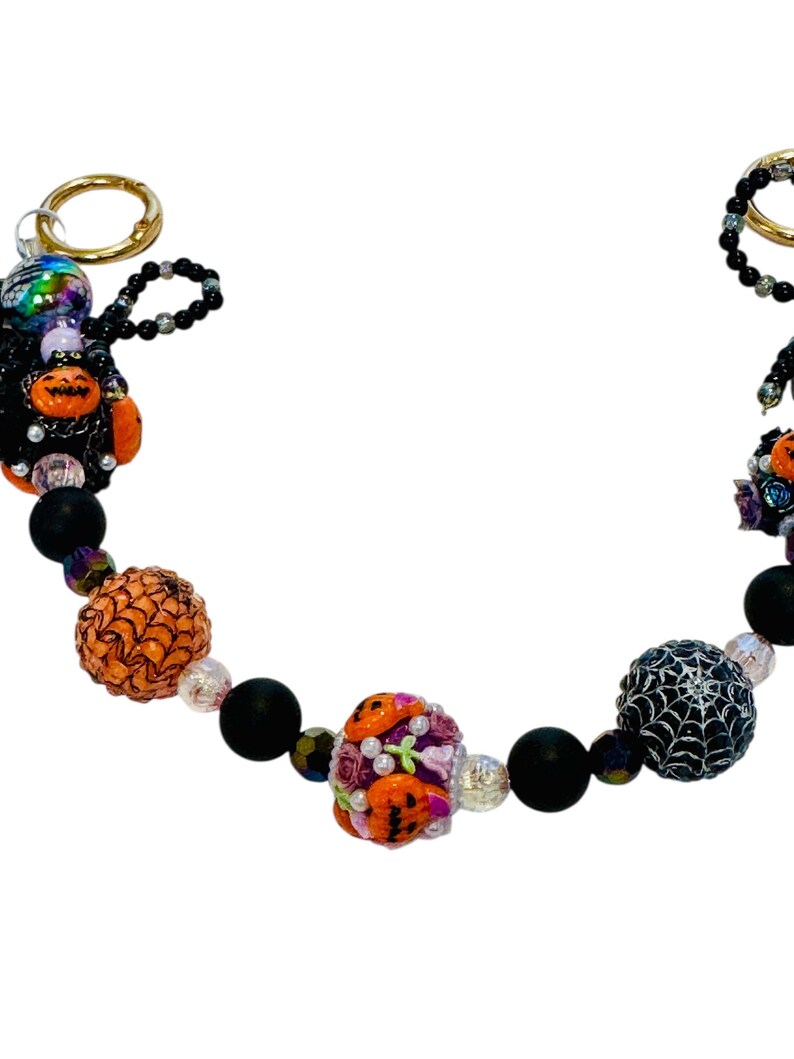 Puede incluir: Un llavero de cuentas con tem&aacute;tica de Halloween con anillos dorados. Las cuentas incluyen cuentas con forma de calabaza naranja, cuentas negras mate y una cuenta con un dise&ntilde;o de telara&ntilde;a. El llavero tambi&eacute;n presenta una figura negra con detalles de calabaza naranja.