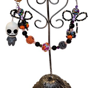 Puede incluir: Llaveros con tem&aacute;tica de Halloween con cuentas decorativas y un dije de Jack Skellington. Los llaveros presentan cuentas negras, naranjas y moradas, con una variedad de texturas y patrones. Los llaveros se exhiben en un soporte de metal decorativo.