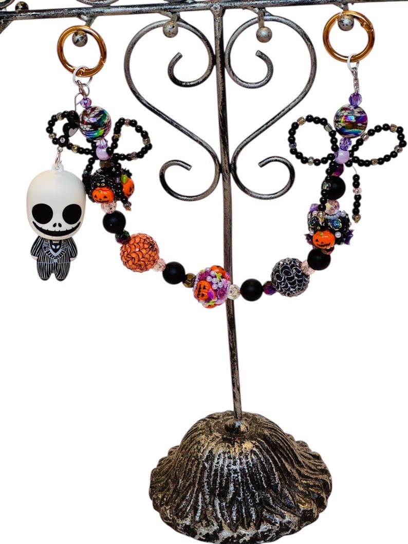 Puede incluir: Llaveros con tem&aacute;tica de Halloween con cuentas decorativas y un dije de Jack Skellington. Los llaveros presentan cuentas negras, naranjas y moradas, con una variedad de texturas y patrones. Los llaveros se exhiben en un soporte de metal decorativo.