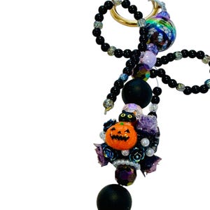 Puede incluir: Un llavero con tem&aacute;tica de Halloween con un anillo dorado, cuentas negras y un grupo decorativo. El grupo incluye una calabaza naranja, un gato negro, rosas moradas y cuentas iridiscentes. El llavero tiene un dise&ntilde;o caprichoso y festivo.