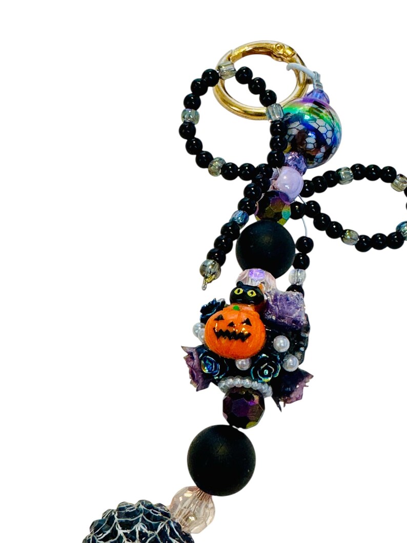 Puede incluir: Un llavero con tem&aacute;tica de Halloween con un anillo dorado, cuentas negras y un grupo decorativo. El grupo incluye una calabaza naranja, un gato negro, rosas moradas y cuentas iridiscentes. El llavero tiene un dise&ntilde;o caprichoso y festivo.