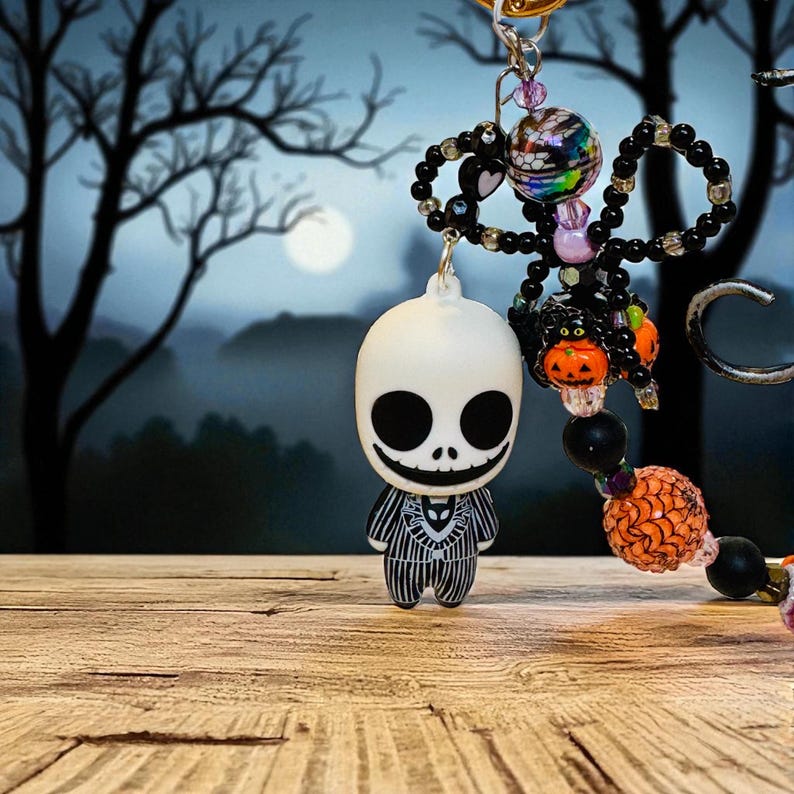Puede incluir: Un llavero tem&aacute;tico de Halloween con una figura blanca de Jack Skellington con un traje a rayas. El llavero incluye cuentas negras, dijes en forma de coraz&oacute;n y detalles de calabaza naranja. El fondo muestra una escena de bosque espeluznante con luna llena.