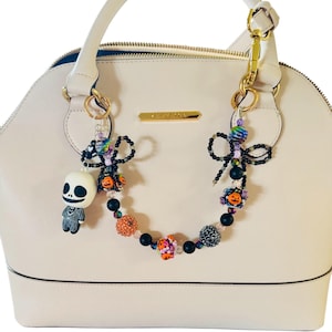 Puede incluir: Un bolso de mano color crema con un charm decorativo de cuentas. El charm presenta una figura de Jack Skellington, cuentas negras y cuentas de calabaza naranja. El bolso tiene un asa superior y herrajes dorados.