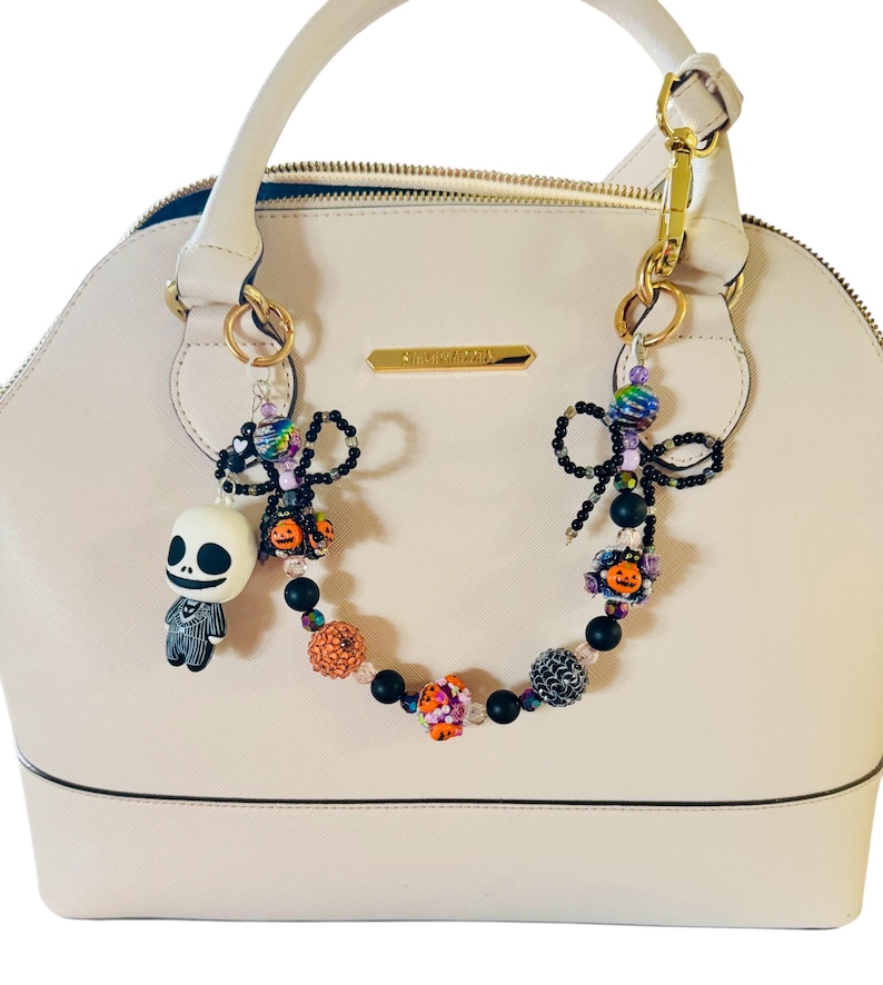 Puede incluir: Un bolso de mano color crema con un charm decorativo de cuentas. El charm presenta una figura de Jack Skellington, cuentas negras y cuentas de calabaza naranja. El bolso tiene un asa superior y herrajes dorados.