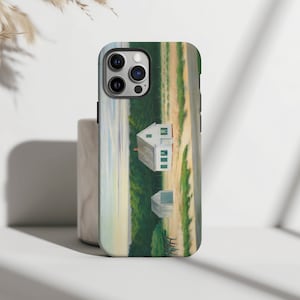 May include: A phone case with a painting of two white houses with green trim on a grassy hill. The sky is a light blue with a hint of orange in the distance.