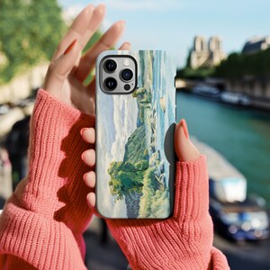 May include: A phone case with a scenic landscape painting of a river and trees. The case has a camera lens cutout and a white background.