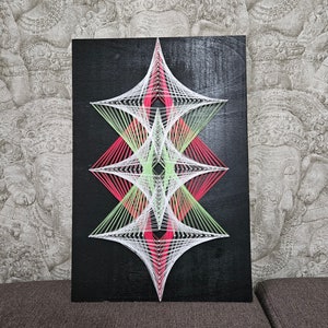 Könnte beinhalten: Ein geometrisches String-Art-Werk mit schwarzem Hintergrund. Das Design besteht aus weißen, rosa und grünen Fäden, die ein symmetrisches Muster bilden.