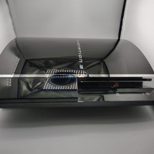 May include: A black PlayStation 3 game console featuring a glossy finish, a silver horizontal trim, and a disc slot on the front. The top surface displays the PlayStation 3 logo in silver lettering, with power and eject buttons located on the right side.