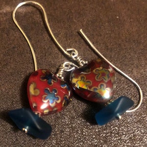 May include: A pair of silver dangle earrings with red heart-shaped glass beads. The beads have a floral pattern and iridescent finish. The earrings are accented with blue glass beads.