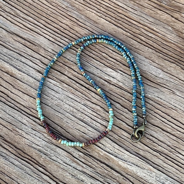 Op de afbeelding: Een veelkleurige kralenketting met een bronzen sluiting. De kralen zijn in een verloop van blauw, groen en bruin.