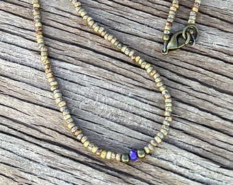 May include: A beaded necklace with small, round, golden-colored beads. The necklace has a bronze-colored clasp and a single, small, purple bead. The necklace is displayed on a wooden surface.