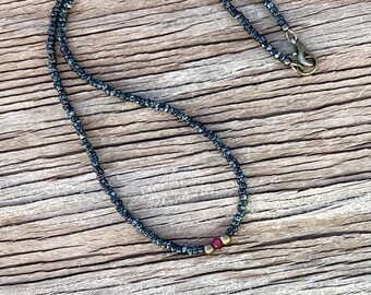May include: A delicate necklace made of small black beads with a single red bead and two gold beads as accents. The necklace is on a wooden surface.