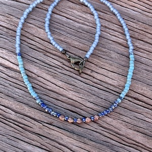 May include: A delicate necklace with a gradient of blue beads, transitioning from light to dark blue, with a few copper beads at the bottom. The necklace has a simple clasp.