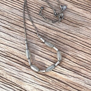 May include: A silver chain necklace with a silver clasp and a tag that says "PD". The necklace has a row of oval-shaped grey beads.