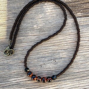 May include: A dark brown beaded necklace with a clasp. The necklace features a series of small, dark brown beads and a section of larger, amber-colored beads with black accents. The necklace is displayed on a wooden surface.