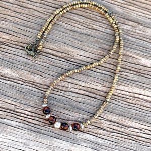 May include: A beaded necklace with a bronze-colored chain and a clasp. The necklace features a series of dark red and white beads, interspersed with smaller silver-colored beads. The necklace is displayed on a wooden surface.