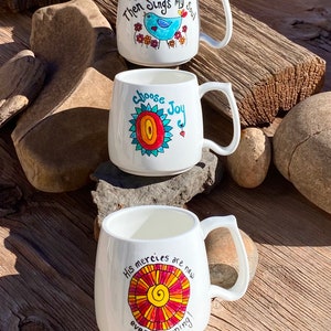 May include: Three white ceramic mugs with colourful hand-painted designs and inspirational quotes. The mugs feature a blue bird, a red and yellow sun, and a red and orange sun. The quotes are "Then Sings My Soul", "Choose Joy", and "His Mercies Are New Every Morning".