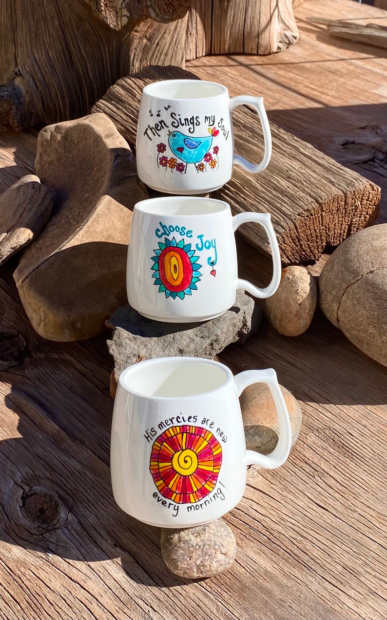 May include: Three white ceramic mugs with colourful hand-painted designs and inspirational quotes. The mugs feature a blue bird, a red and yellow sun, and a red and orange sun. The quotes are "Then Sings My Soul", "Choose Joy", and "His Mercies Are New Every Morning".