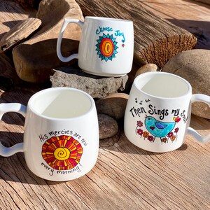 May include: Three white ceramic mugs with colourful hand-painted designs. The mugs feature inspirational quotes and images of a sun, a bird, and flowers. The quotes are "Choose Joy", "His mercies are new every morning", and "Then Sings my Soul".