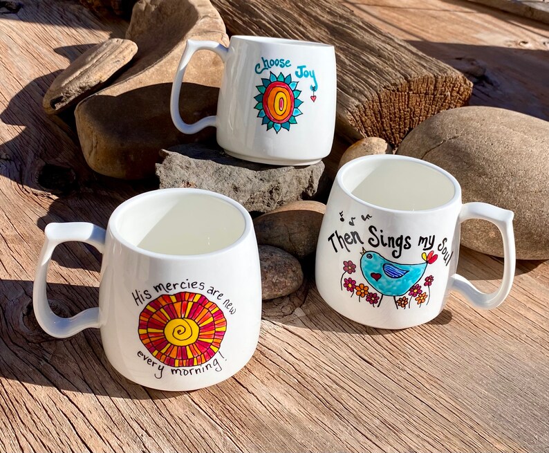May include: Three white ceramic mugs with colourful hand-painted designs. The mugs feature inspirational quotes and images of a sun, a bird, and flowers. The quotes are "Choose Joy", "His mercies are new every morning", and "Then Sings my Soul".