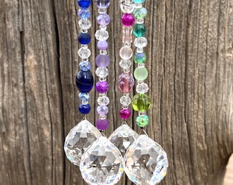 May include: Four crystal suncatchers with beaded strands in various colors, including blue, purple, pink, and green. The strands are hanging from a wooden surface.