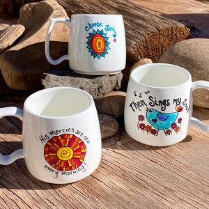May include: Three white ceramic mugs with inspirational quotes and colourful illustrations. The mugs feature a sun with the words "Choose Joy", a bird with the words "Then Sings My Soul", and a sun with the words "His mercies are new every morning!"