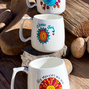 May include: Three white ceramic mugs with colourful hand-painted designs. The first mug features a blue bird with the text "Then Sings my Soul". The second mug features a colourful sun with the text "Choose Joy". The third mug features a colourful sun with the text "His mercies are new every morning!"