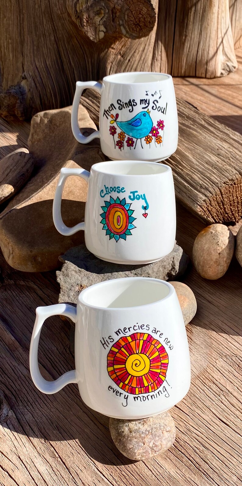May include: Three white ceramic mugs with colourful hand-painted designs. The first mug features a blue bird with the text "Then Sings my Soul". The second mug features a colourful sun with the text "Choose Joy". The third mug features a colourful sun with the text "His mercies are new every morning!"