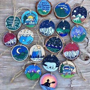 May include: A collection of 14 round wood slice ornaments painted with various mountain scenes, stars, and inspirational quotes. The ornaments are decorated with blue, green, red, white, and black paint. Some of the quotes include "I yearn for mountains that I've never even seen", "Adventure awaits", "Life is good", and "The mountains are calling".