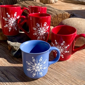 May include: A collection of ceramic mugs in red and blue, each adorned with a white snowflake design. The mugs have curved handles and are arranged on a wooden surface, perfect for enjoying hot beverages.