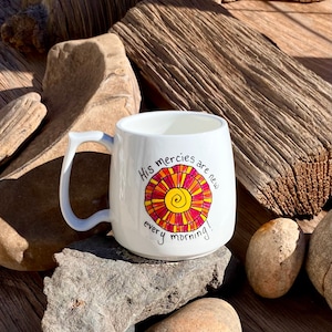 May include: White ceramic mug with a colourful sun design and the text "His mercies are new every morning!"