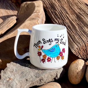 May include: White ceramic mug featuring a blue bird singing with the text "Then Sings my Soul" and musical notes. The bird is surrounded by colourful flowers.