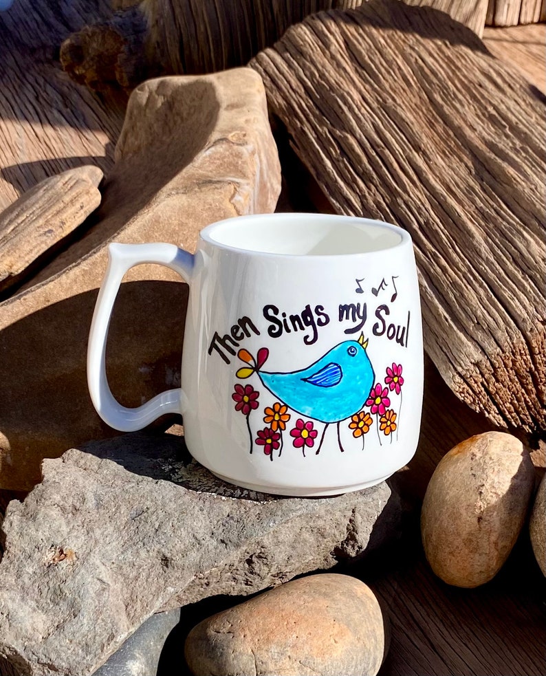 May include: White ceramic mug featuring a blue bird singing with the text "Then Sings my Soul" and musical notes. The bird is surrounded by colourful flowers.