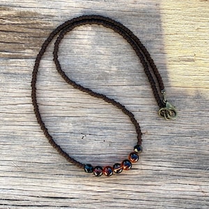 May include: A brown beaded necklace with a clasp. The necklace features small, dark brown beads and larger, round, amber-colored beads. The clasp is a bronze color. The necklace is displayed on a wooden surface.