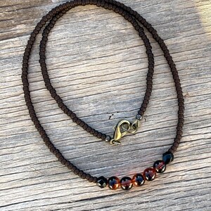 May include: A brown beaded necklace with a clasp. The necklace features small, dark brown beads and a row of larger, amber-colored beads. The clasp is a bronze color. The necklace is displayed on a wooden surface.