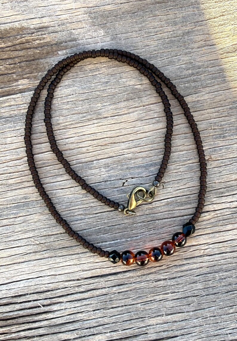 May include: A brown beaded necklace with a clasp. The necklace features small, dark brown beads and a row of larger, amber-colored beads. The clasp is a bronze color. The necklace is displayed on a wooden surface.